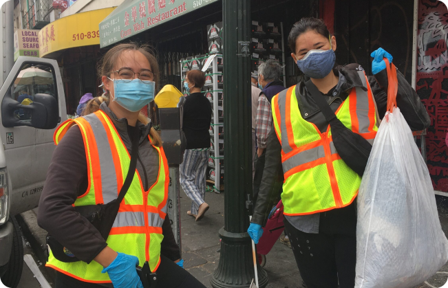 Two APEN volunteers standing on a street in Oakland Chinatown wearing clean-up gear with face masks, hand gloves, trash bag, and trash picker