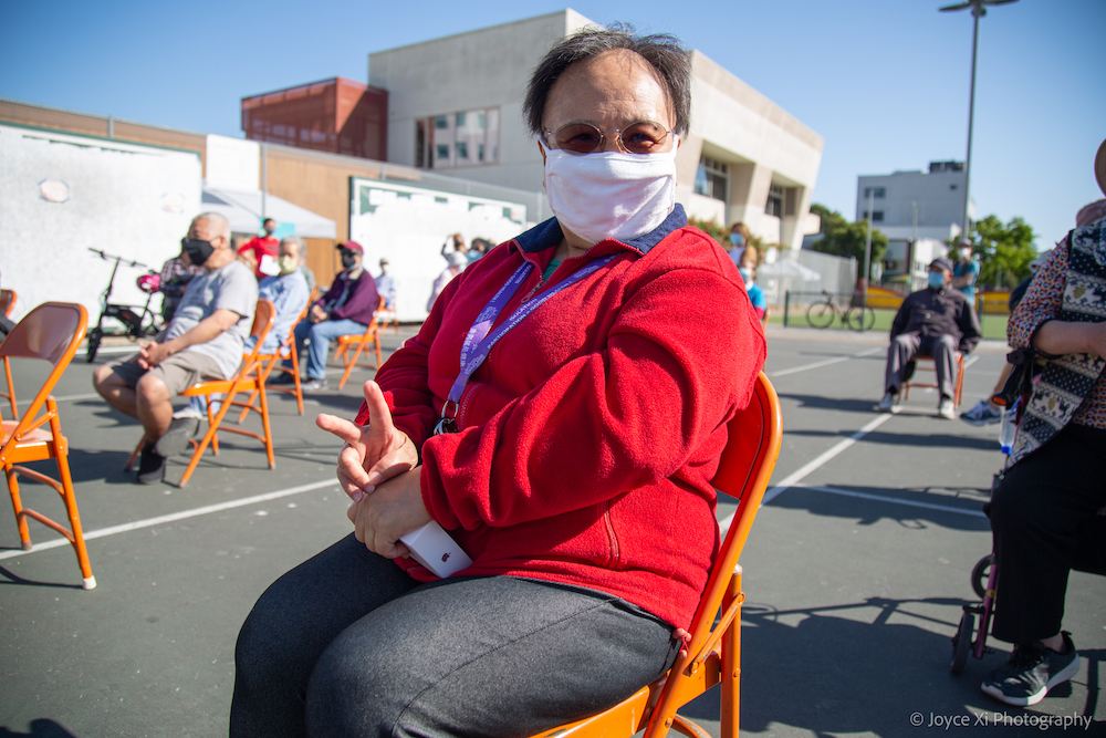 An APEN Oakland Chinatown member sits in a chair on a concrete ground in a park, making a peace sign at the camera and wearing a mask
