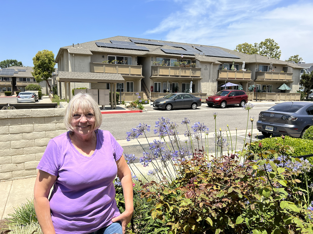 A woman in a purple shirt smiles at the camera standing in front of an apartment building.