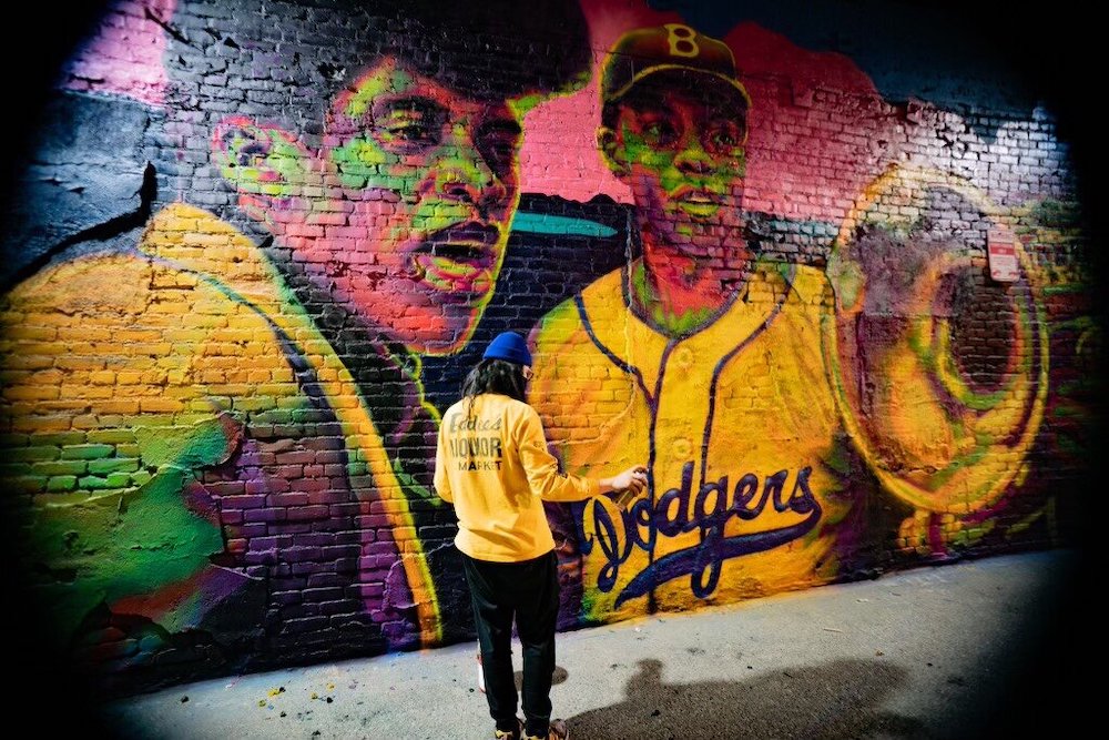 A person in a yellow shirt with long hair faces away from the camera, in front of a mural that includes Jackie Robinson.