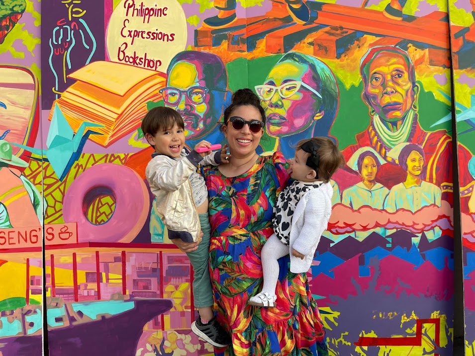 Aysha Pamukcu stands holding her two kids in front of a colorful mural smiling at the camera